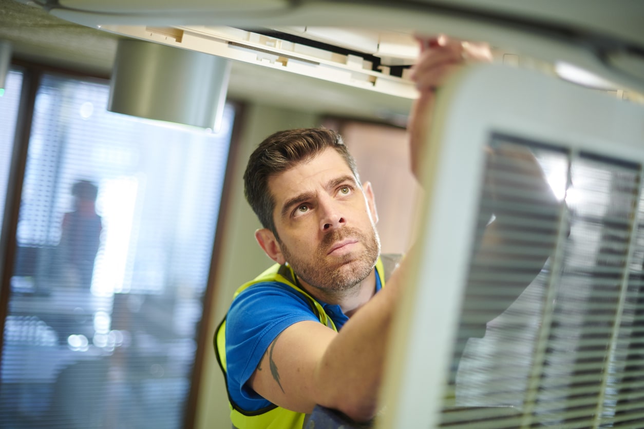 A man in a safety vest repairs an air conditioner on a building rooftop.