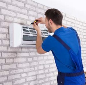 A man in overalls repairs an air conditioner, focused on the unit with tools in hand.